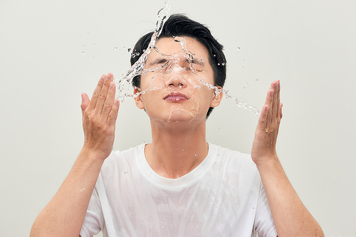 Handsome young man is washing his face, splashes of water around, white background