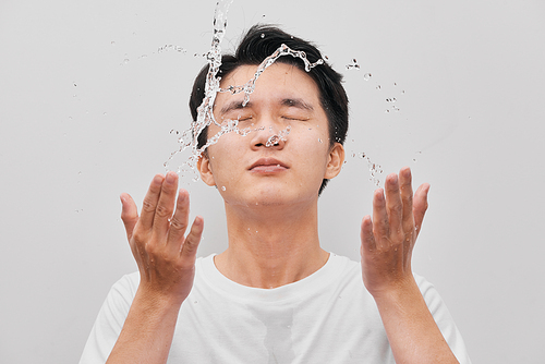Young man spraying water on his face over gray background
