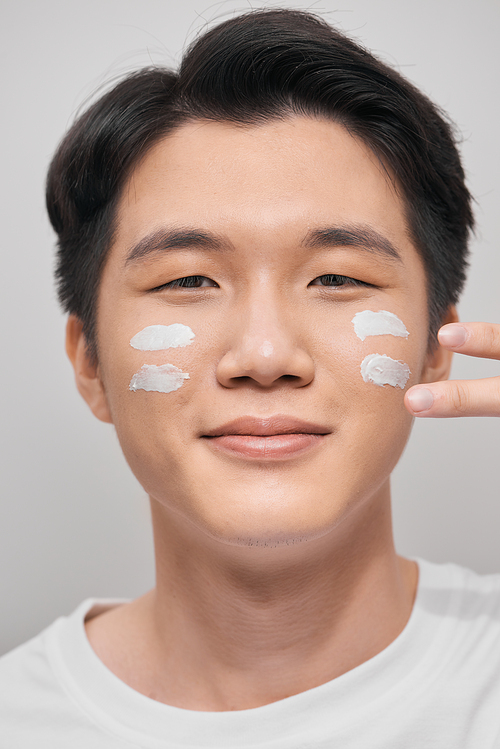 Skin care. Portrait of handsome young man applying cream to his face, isolated on white background