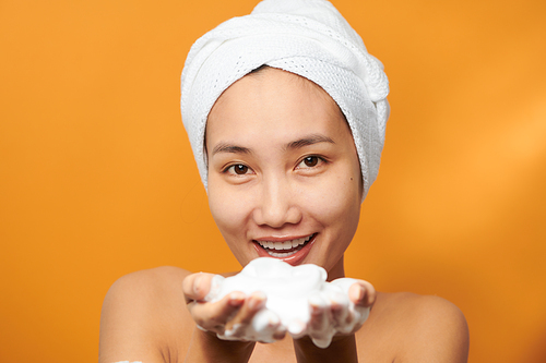 Young Asian woman showing foaming cleanser on her hand while wearing a bath towel over orange background.