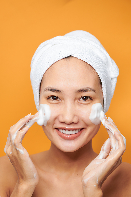 Happy young Asian woman applying face cream while wearing a towel and touching her face. Isolated on orange background