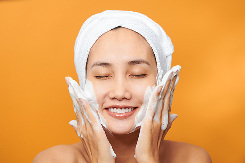 Happy young Asian woman applying face cream while wearing a towel and touching her face. Isolated on orange background
