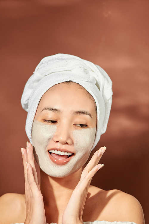 Close-up portrait of attractive girl with a towel on head and clay mask on face isolated over orange background.
