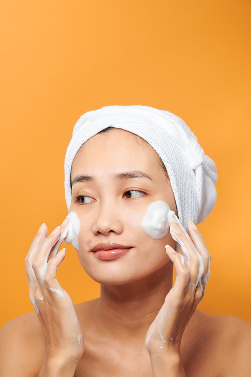Close up beauty portrait of a laughing beautiful half naked Asian woman applying face cream and looking away isolated over orange background