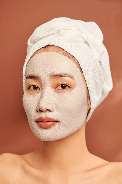 Portrait of young Asian woman on orange background with clay mask on her face and a towel on her head smiling.