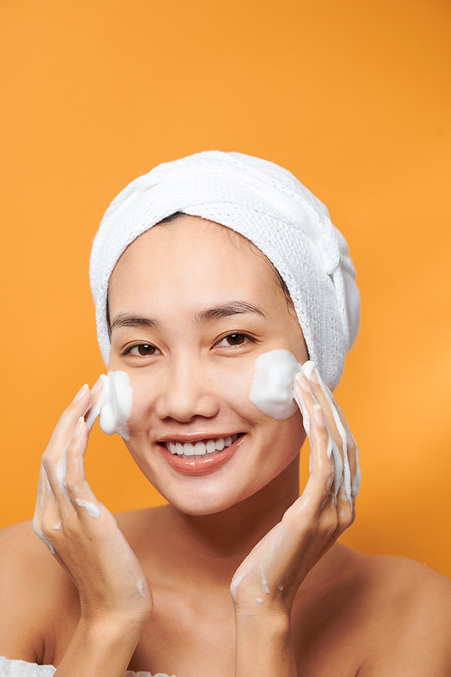 Young Asian woman applying moisturizing cream on her face. Photo of woman towel on orange background. Skin care concept