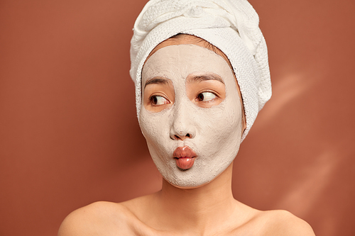 Spa girl with pleased facial expression, applies clay mask on face, gets beauty treatments, wears white soft towel on head