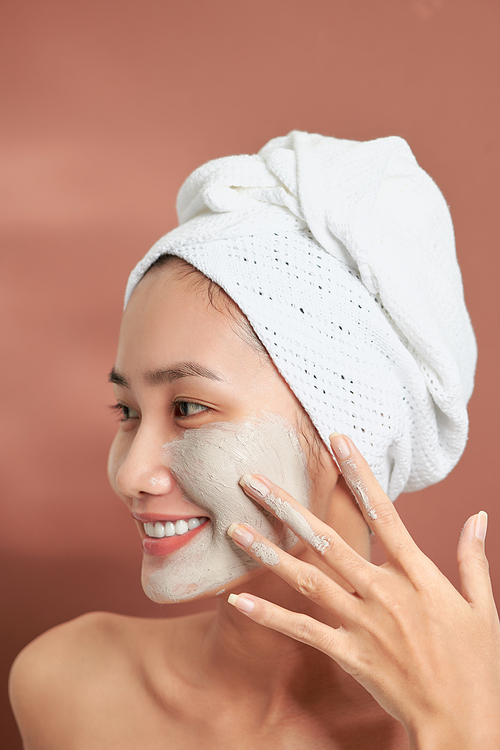 Beautiful cheerful Asian teen girl applying facial clay mask. Beauty treatments, isolated on light background.