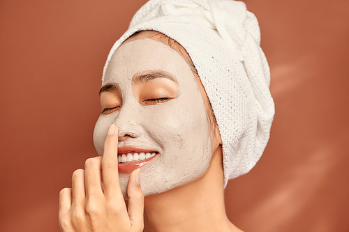 Happy young Asian woman applying clay mask on her face over orange background. Close up.