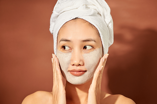Portrait of young Asian woman on orange background with clay mask on her face and a towel on her head smiling.