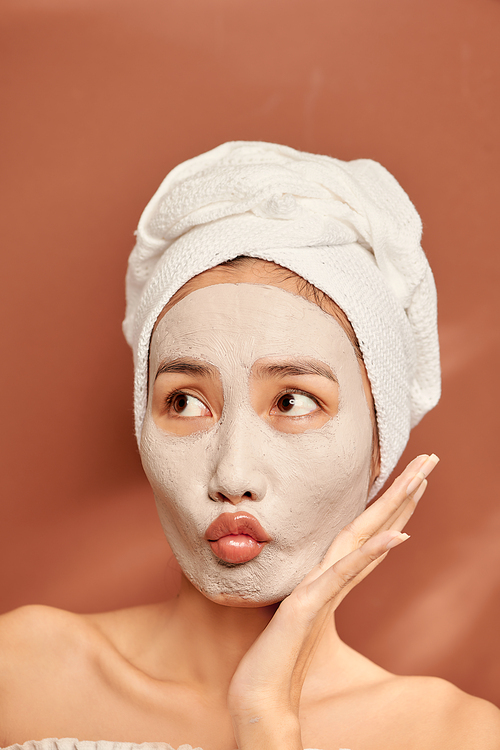 Spa girl with pleased facial expression, applies clay mask on face, gets beauty treatments, wears white soft towel on head