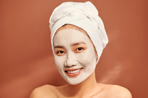 Close-up portrait of attractive girl with a towel on head and clay mask on face isolated over orange background.