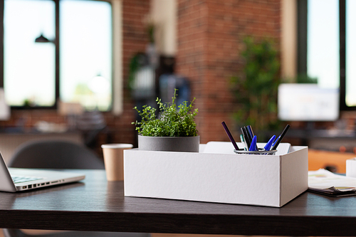 Close up of office supplies in box on wooden desk. Houseplant in pot, decorations and laptop on table in empty business space. Flowerpot, modern ornaments and computer at work