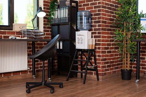 Close up of bookshelf and water dispenser in business office. Empty desk with decorations and watercooler in startup space, shelves with minimal decor. Decorative plants and refreshment.