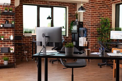 Empty startup office with computer and supplies on desk. Nobody in business space used to work on company growth with monitor and equipment. No people in workplace with decorations.