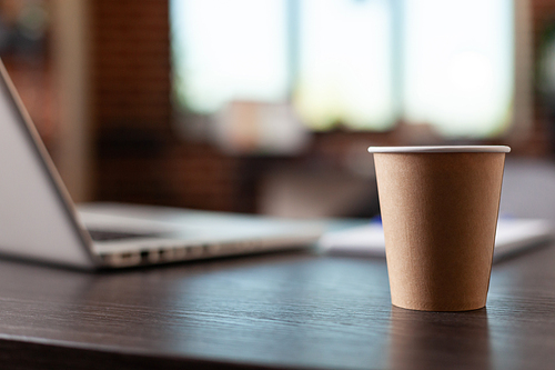 Close up of disposable coffee cup and laptop on office desk in startup space. Hot beverage on table in empty business workplace. Drink in plastic cup on furniture and computer.