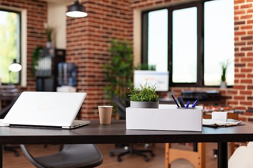 Close up of laptop and box with office supplies on desk. Empty startup office with modern computer and decorations on wooden table. Nobody in workspace with plant and instruments