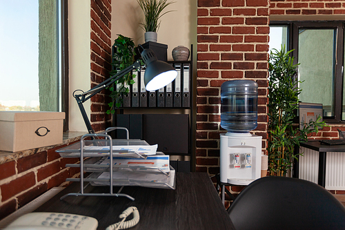 Close up of office corner with desk and bookshelf in empty startup space. Nobody at wooden table with computer and supplies to work on business, shelves with modern decorations.