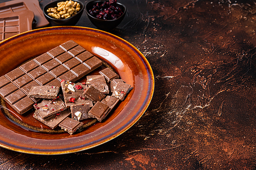 Homemade milk chocolate with hazelnuts, peanuts, cranberries and freeze dried raspberries on a rustic plate. Dark background. Top view. Copy space.