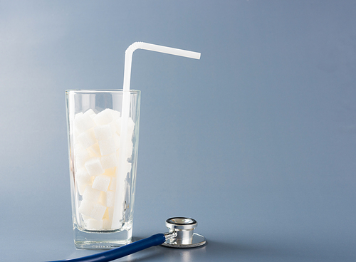 A glass full of white sugar cube sweet food ingredient and doctor stethoscope, studio shot isolated gray background, health high blood risk of diabetes and calorie intake concept and unhealthy drink