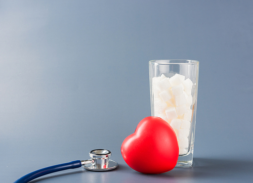 Red heart and glass full of white sugar cube sweet food ingredient and doctor stethoscope isolated on gray background, health high blood risk of diabetes and calorie intake concept and unhealthy drink