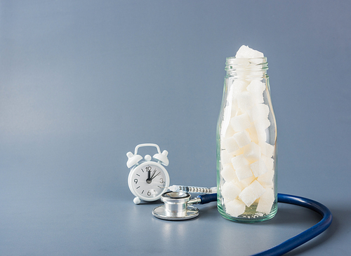 Glass bottle full of white sugar cube sweet food ingredient and doctor stethoscope isolated gray background, health high blood risk of diabetes and calorie intake concept and unhealthy drink