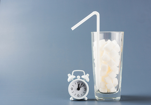 A glass full of white sugar cube sweet food ingredient and alarm clock, studio shot isolated on a gray background, health high blood risk of diabetes and calorie intake concept and unhealthy drink