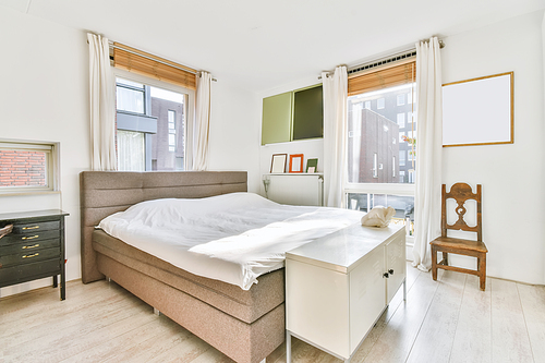 Sunlit bed covered with blanket in light contemporary bedroom with shelves and decorations