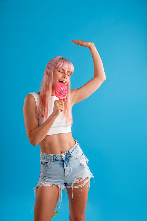 Lively woman with pink hair having fun, holding hair comb and singing in it while standing isolated over blue studio background. Beauty, hair care, lifestyle concept