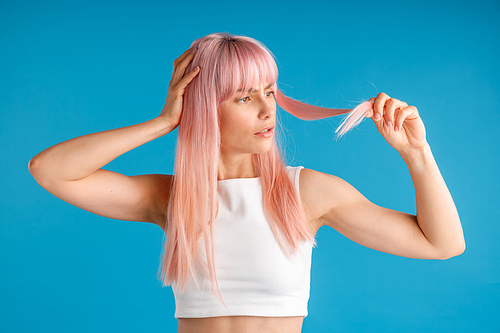 Confused young woman holding and looking at her natural long pink dyed hair while posing isolated over blue studio background. Beauty, hair care concept