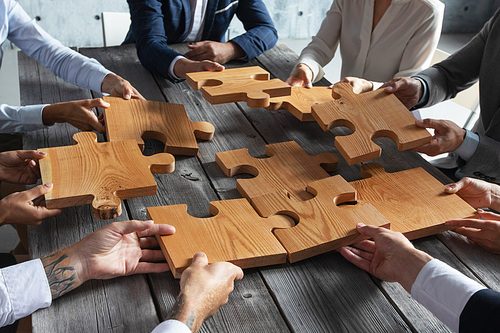 Business people team sitting around meeting table and assembling wooden jigsaw puzzle pieces unity cooperation ideas concept