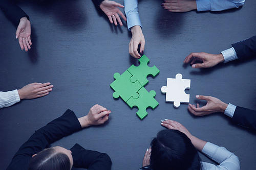 Group of business people assembling jigsaw puzzle at office table, top view, team support and help concept, green and white pieces