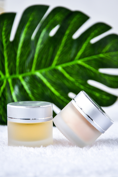 Glass jar with cream organic body care cosmetic products on monstera palm leaves over white textile background. SPA branding mock-up. Moisturizing care skincare face cream for healing complicated troubled skin type