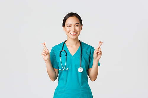 Covid-19, healthcare workers, pandemic concept. Optimistic smiling asian female doctor, physician in scrubs having faith, cross fingers good luck, making wish, hope for better, white background.