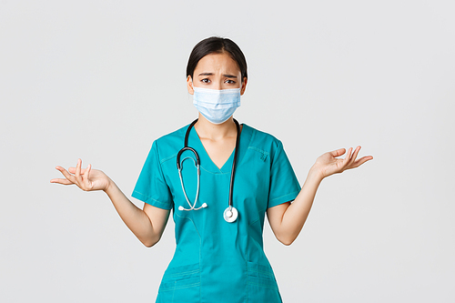 Covid-19, coronavirus disease, healthcare workers concept. Troubled and confused asian female nurse in scrubs and medical mask asking question, shrugging and look concerned, white background.