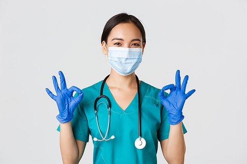 Covid-19, coronavirus disease, healthcare workers concept. Confident smiling asian female doctor, nurse in medical mask and gloves, show okay gesture in approval, white background.