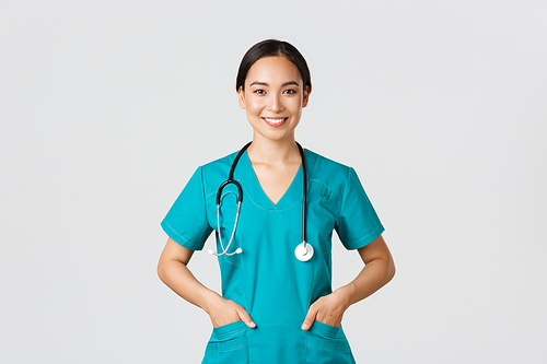 Covid-19, healthcare workers, pandemic concept. Confident smiling pretty asian female doctor, physician looking determined and upbeat, holding hands in pockets of scrubs, examine patients in clinic.