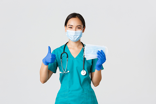Covid-19, coronavirus disease, healthcare workers concept. Smiling asian doctor, nurse in scrubs and rubber gloves, showing thumbs-up and medical masks, white background.