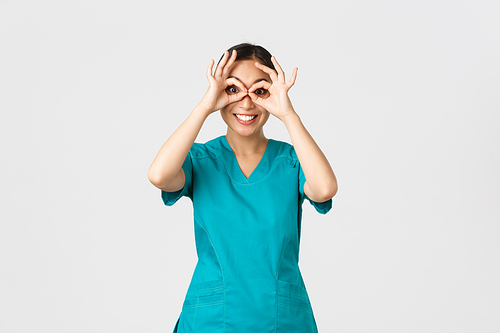 Covid-19, healthcare workers and preventing virus concept. Amused happy, cute asian female doctor, intern in scrubs looking through hand binoculars with excited expression, smiling.