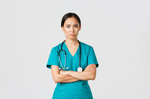Covid-19, healthcare workers, pandemic concept. Skeptical and reluctant asian female doctor, tired nurse in scrubs cross arms and frowning, smirk displeased, standing white background.