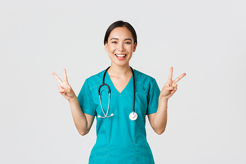 Covid-19, healthcare workers, pandemic concept. Happy asian female doctor, nurse in scrubs showing peace kawaii gesture and smiling, staying positive during work in hospital, white background.