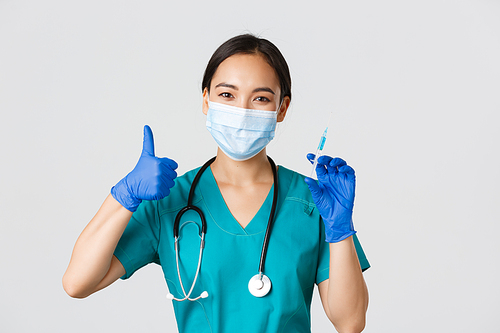 Covid-19, coronavirus disease, healthcare workers concept. Smiling beautiful asian medical worker, nurse in mask and gloves thumb-up, hold syringe with vaccine, standing white background.