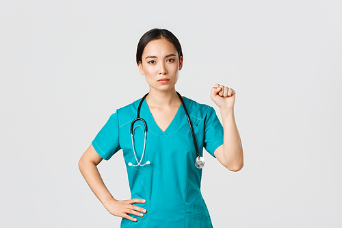 Covid-19, healthcare workers, pandemic concept. Serious-looking confident serious asian female doctor showing support to fellow collegues during coronavirus, raising fist in unity gesture.
