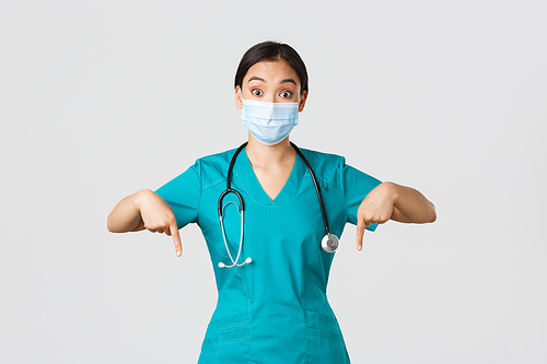 Covid-19, coronavirus disease, healthcare workers concept. Excited and surprised asian female physician, nurse in scrubs and medical mask, pointing fingers down, showing advertisement.