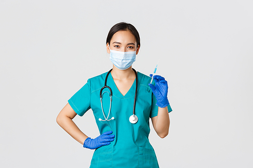 Covid-19, coronavirus disease, healthcare workers concept. Smiling confident asian female nurse in scrubs and medical mask, holding syringe with vaccine, doing vaccination shot, white background.