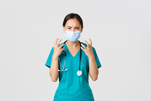 Covid-19, coronavirus disease, healthcare workers concept. Angry and pissed-off asian female doctor, physician in medical mask and scrubs, clench fists outraged, looking furious, white background.