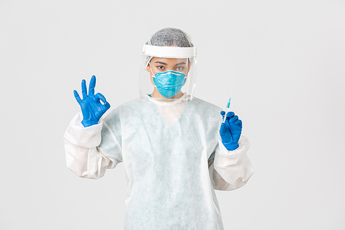 Covid-19, coronavirus disease, healthcare workers concept. Confident serious female asian doctor in personal protective equipment, show okay gesture, hold syringe with vaccine, white background.