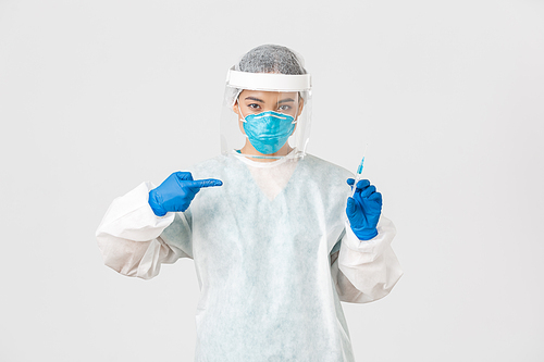 Covid-19, coronavirus disease, healthcare workers concept. Serious asian female doctor, physician in PPE personal protective equipment, pointing finger at syringe with vaccine, white background.