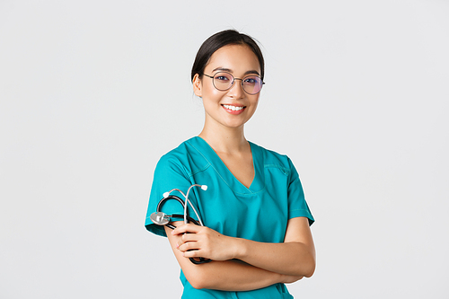 Covid-19, coronavirus disease, healthcare workers concept. Professional good-looking asian doctor, medical worker in glasses and scrubs, cross arms and smiling, white background.