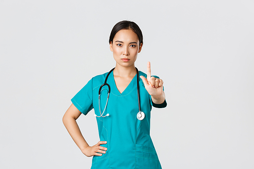 Covid-19, healthcare workers, pandemic concept. Serious-looking professional female asian doctor in scrubs, physician shaking finger in prohibition, warning gesture, forbid something.
